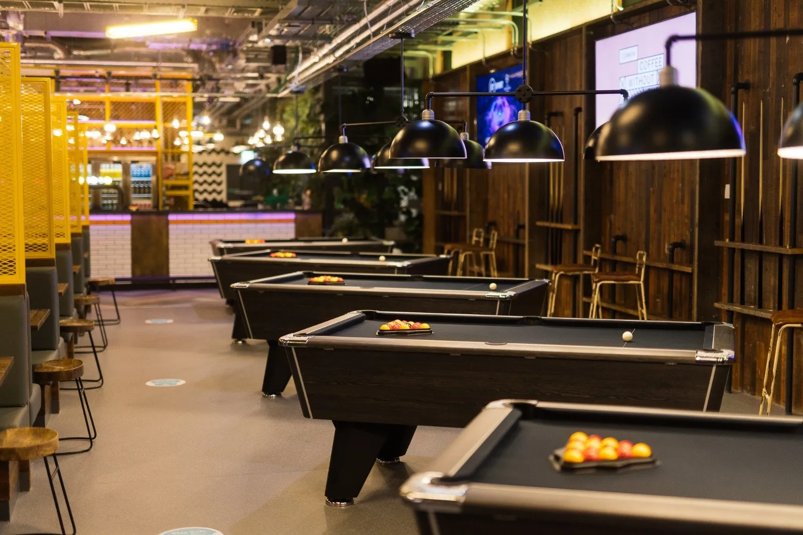 This vibrant, modern entertainment space features a row of dark wood pool tables with black felt, each illuminated by a series of black dome pendant lights. To the left, a lively bar area with white subway tiles and a purple LED accent is flanked by grey upholstered booth seating, separated by striking yellow perforated metal screens. The right wall showcases rustic reclaimed wood paneling, while the exposed industrial ceiling and polished concrete floor contribute to a dynamic, urban atmosphere perfect for social gatherings.