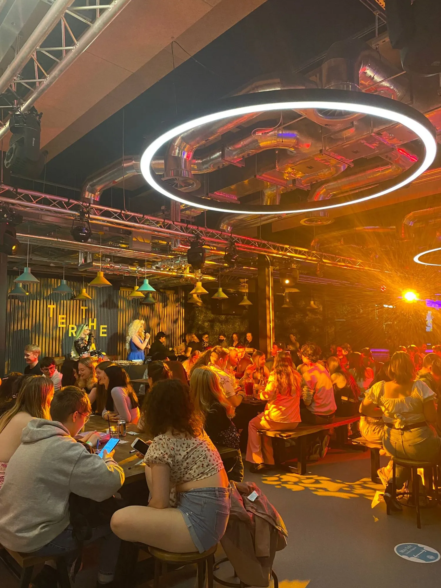 This vibrant interior shot captures a bustling, industrial-chic hospitality venue during an evening event, likely a live performance. The space features high ceilings with exposed metallic ductwork and structural trusses, illuminated by a mix of warm amber stage lighting, cool white circular LED pendants, and colourful industrial-style bell pendants. Guests are seated at long communal wooden tables with benches and stools, creating an energetic and social atmosphere, with a corrugated metal wall serving as a dynamic backdrop for the performers.
