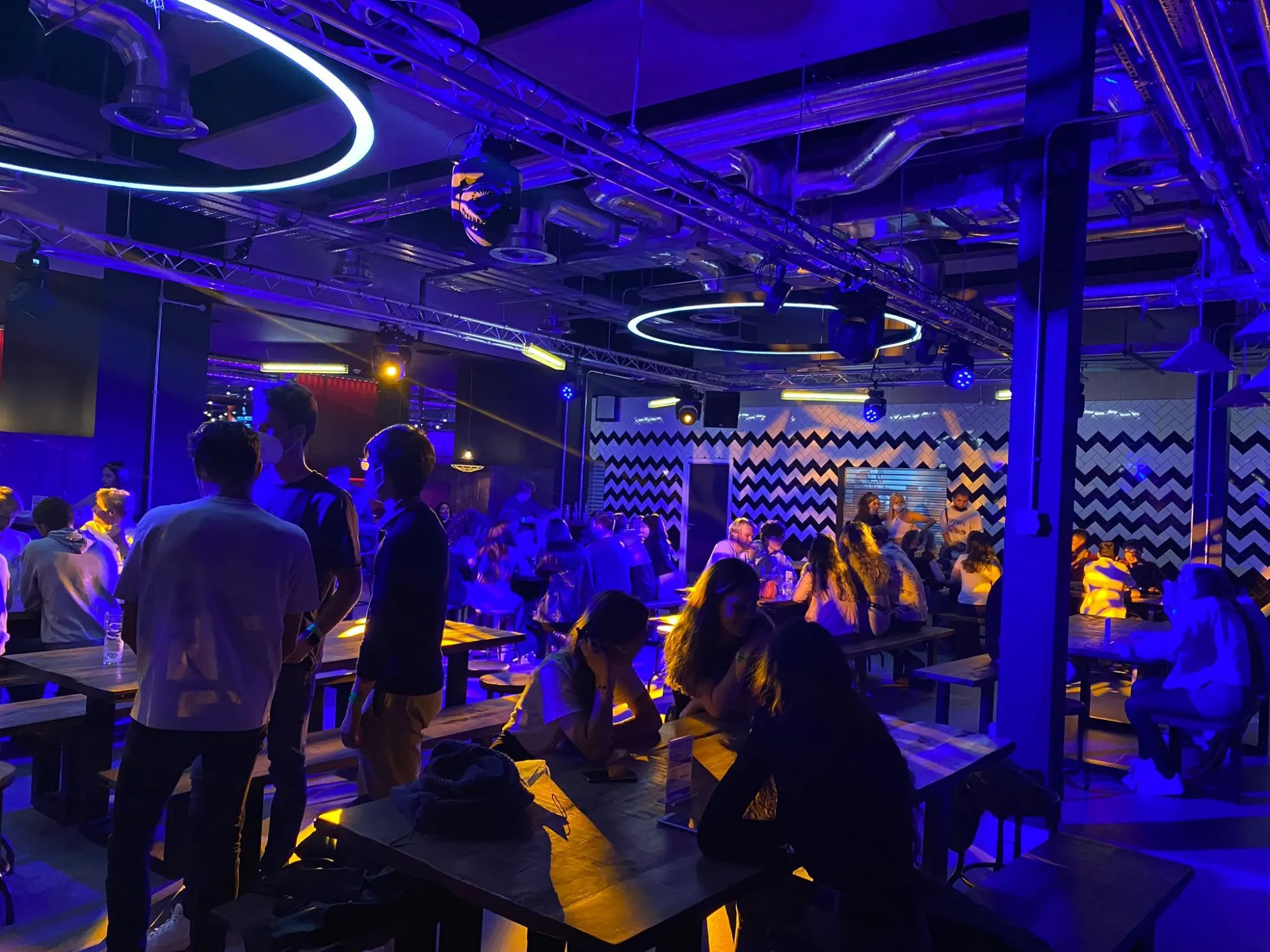 This vibrant, industrial-chic entertainment venue features an open-plan layout with exposed metal trusses and ductwork across the ceiling, creating a raw, urban aesthetic. The space is dramatically lit with a dominant deep blue and purple wash, punctuated by warm yellow spotlights illuminating the rustic wooden communal tables and benches where patrons are socializing. A striking black and white zigzag patterned wall adds a playful, graphic element, contributing to the energetic and modern club-like atmosphere.