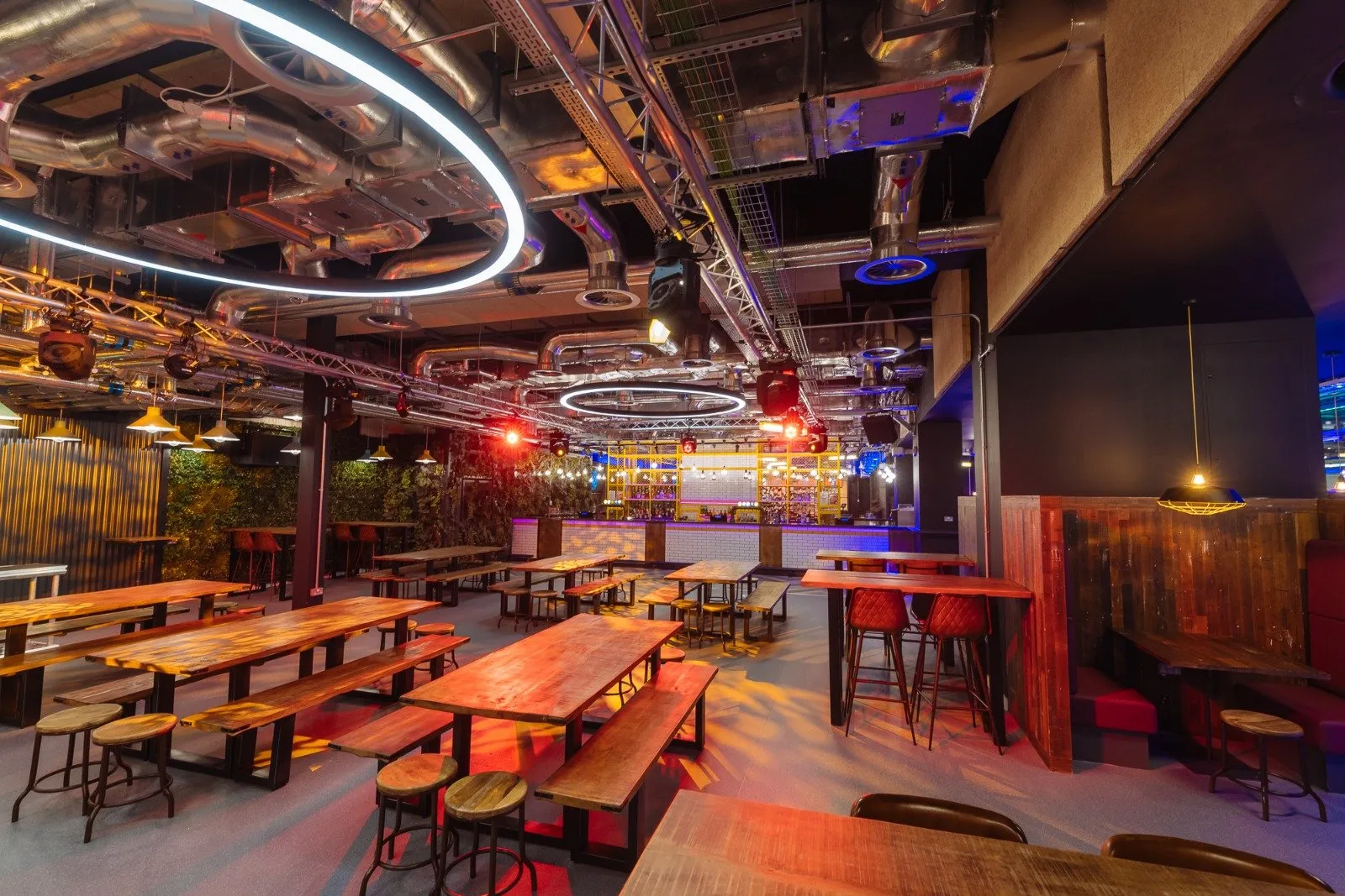 This vibrant, industrial-chic hospitality space features an expansive open-plan layout with a dynamic mix of seating. Exposed galvanized ductwork, trusses, and track lighting dominate the high ceiling, complemented by striking circular neon fixtures and colorful LED washes that create an energetic, club-like atmosphere. A variety of reclaimed wood communal tables with benches and stools, alongside high-top tables and cozy red upholstered booths, offer diverse dining and social zones, all anchored by a brightly lit bar with subway tile and geometric shelving at the rear.