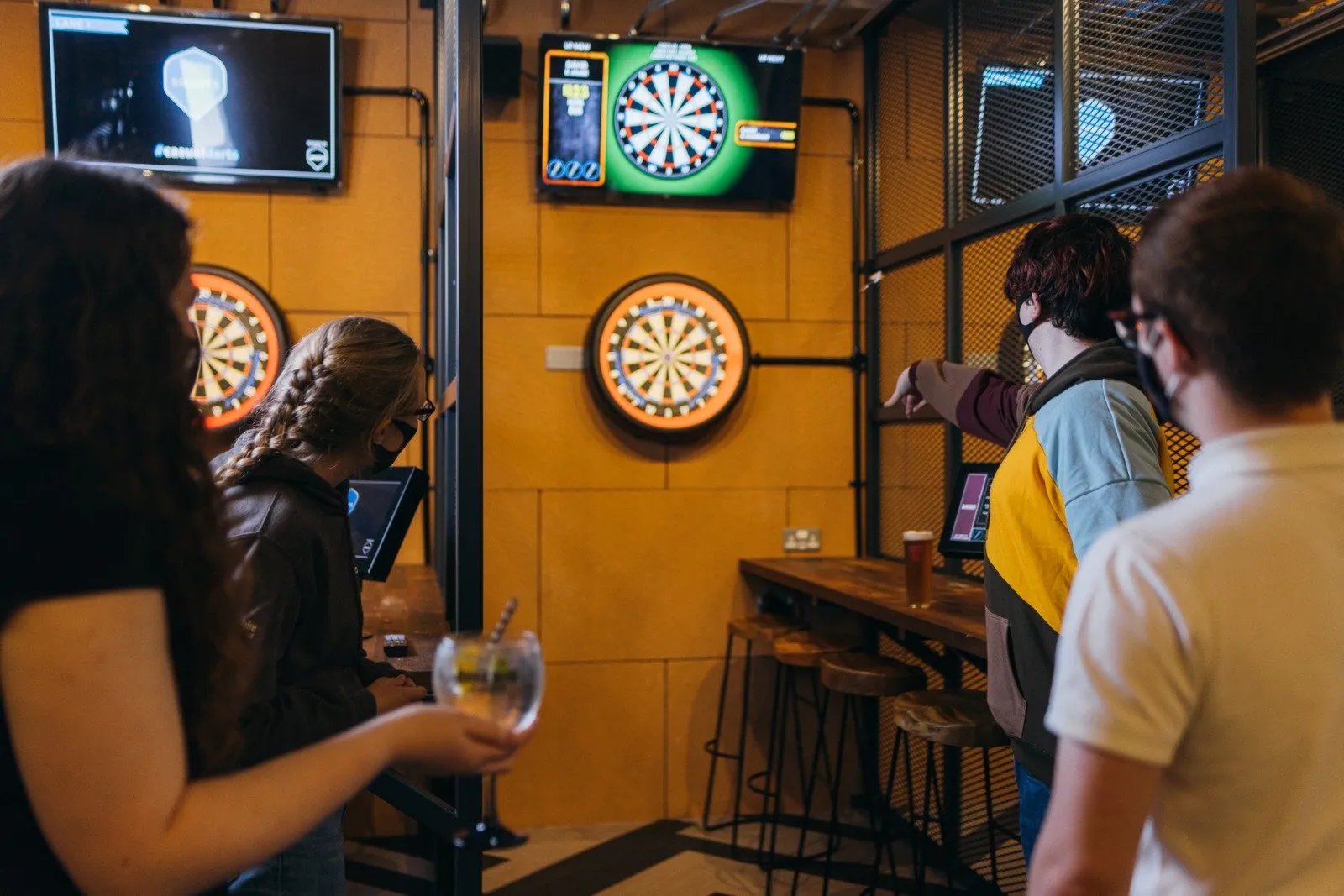This vibrant shot captures a lively, modern entertainment space dedicated to darts. The design features warm, block-patterned wood wall cladding contrasted with industrial black metal mesh partitions and exposed conduit, creating a contemporary yet robust aesthetic. Each dart station is equipped with a backlit dartboard, a digital scoring screen, and a high-top wooden counter with matching backless stools, fostering a social and engaging atmosphere for guests enjoying drinks and games.